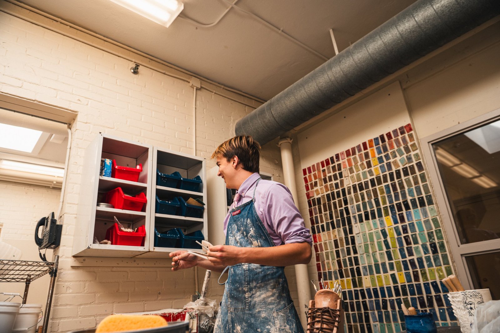 Me in my apron laughing in the ceramics studio, with a wall of glaze test tiles behind me